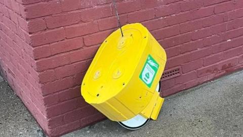 A yellow defibrillator which has been kicked off the wall and as hanging by a wire.