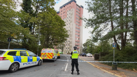 A lone police officer stands in front of a police cordon. A pink and white multi-storey is in the background