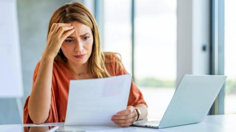 A woman in a loose but smart top looks at papers whilst sitting in front of an open laptop. She is frowning slightly.