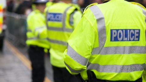 Three police officers standing outside with their backs to the camera. They are all wearing fluorescent yellow jackets with silver stripes along the arms and backs. On the back of each of their jackets in a blue rectangle and in grey writing it says POLICE.