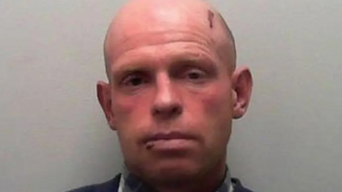 The image shows an upper‑body view of Huw Owen, a bald, red-faced man, standing against a plain, light‑coloured background, in a police mug shot. He is wearing a dark blue sweater over a collared shirt that has a checked pattern.