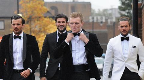 TOWIE stars wearing tuxedos walking along a residential street to Sugar Hut. All tuxedos, completed with bow ties, are black with the exception of Pete Wicks, whose jacket is white. They all appear to be having a laugh together.