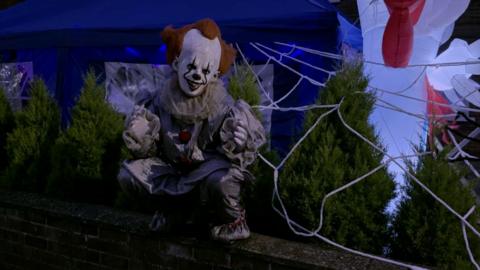 A person in a detailed clown costume is sitting on a brick wall in front of a house decorated with a blue canopy, large white spiderwebs, green shrubs, and an inflatable red-and-white Halloween figure.