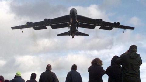 A B52 Stratofortress bomber flying out of RAF Fairford. There is a group of people below watching the plane take off.