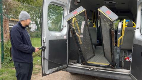 A private hire operator, wearing a wooly hat, uses a handset to lower a lift used to get wheelchairs into the back of a specially adapted minibus.