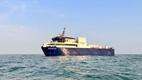 A blue and white freight vessel bobs on the surface of calm waters. The sun is shining on the side of the boat's exterior. 