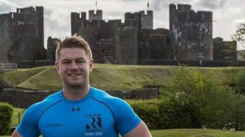 A photo of a man looking at the camera. Behind him is Caerphilly Castle. It is a head and shoulders shot. He has a blue t-shirt on and short blonde hair as he smiles. 