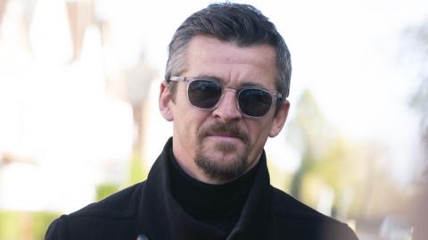 Joey Barton, who has greying hair swept to the right and a goatee beard, looks into the camera wearing a black woollen style coat over a black  turtle-neck jumper. 