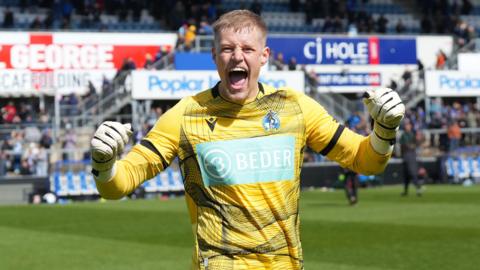 Bristol Rovers goalkeeper Brad Young celebrates with both fists in the air after helping his team to victory in League Two. Young is wearing a yellow goalkeeping jersey and white gloves