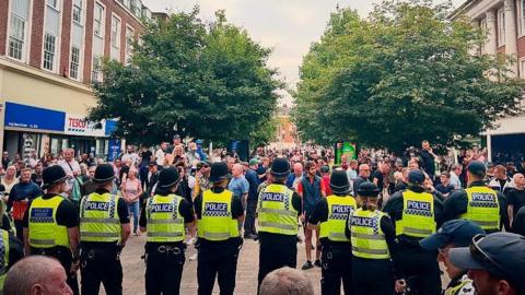 A row of police officers have their back to the camera as they are approached by a large crowd in Hull city centre. The officers are wearing high-visibility vests and police helmets. Two rows of shops flank each side of the street which also has large trees.