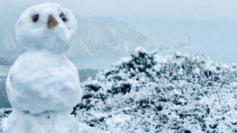 A snowy scene from a clifftop above a beach. A small snowman has been made with three items places on its head to make eyes and a nose.