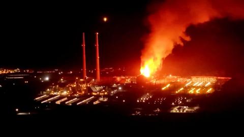 A fire burns at night at a power plant beside two tall chimneys