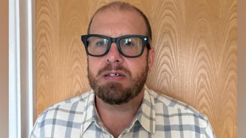 A man wearing smart glasses. He has short hair and a thick beard. He is also wearing a white and blue chequered shirt. There is a wooden door behind him.