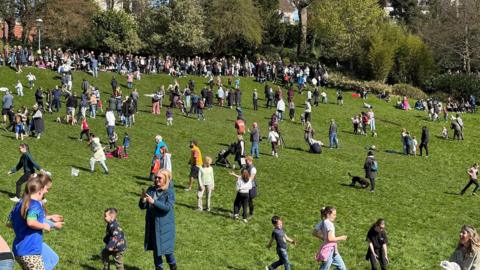 Large crowds of people of all ages are scattered over the large grassy slope with a line of families watching from the top ridge with trees behind.