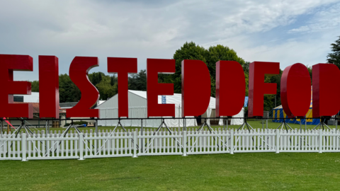 Big red letters spelling out the word Eisteddfod, they are at the centre of the image, with white picket fences in front of the letters. A white tent is visible just behind and there is grass in front of the picture, showing the image is taken on a field.
