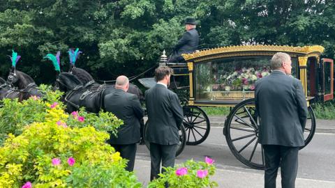 A row of three men in suits watching a horse-drawn carriage pass for a funeral procession. There is a coffin draped in flowers in the carriage, which is being led by black horses with feather headdresses.