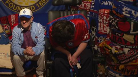 A man sits next to a younger boy in a wheelchair with a wall of football scarves behind 