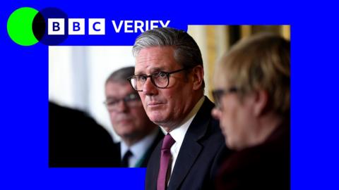 Sir Keir Starmer speaking in a meeting. He is wearing a dark suit with glasses and two other people sit on either side of him. The BBC Verify colours and logo are imposed over it. 