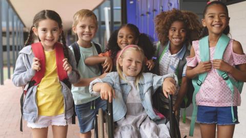 six kids in a school backdrop outside all smiling and with backpacks.