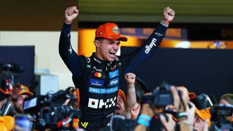 A racing driver dressed in a black racing suit with sponsors' badges all over the front. He has his hands aloft and is being supported on the shoulders of those in  crowd. 