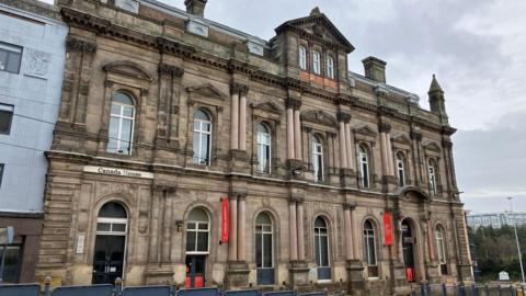 Large impressive Victorian light stone, three storey building in the centre of the image. The building has for visible red banners attached to two of its doors and partway up the wall by its second and fifth window frames.
In the foreground are a series nine square, solid blue metal protective barriers, transitioning into a series of see through blue barriers, a demarcation for trams. 