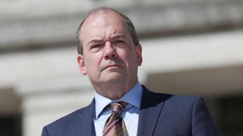 Michael McBride is looking down at the camera. He is unsmiling, with short thinning grey hair, and he's wearing a blue shirt and suit with a multi-coloured tie. 
