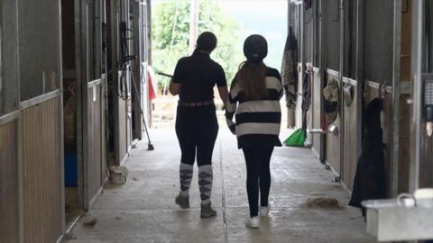 Two young girls wearing riding hats and with their backs to the camera walk between two rows of stables. 