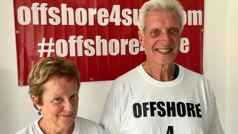 Two people wearing white T-shirts with the slogan "Offshore 4 Sure" are standing in front of a red campaign banner.