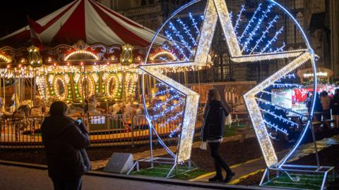 The Christmas Fair 2024 in Canterbury Cathedral precinct. A woman taking a photo of another woman in front of a giant illuminated star with fairground rides in the background