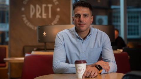 Pret A Manger boss Pano Christou, wearing a blue shirt, sits in a Pret store with a takeaway coffee cup in front of him.
