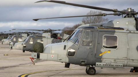 A row of Wildcat helicopters, painted with grey camoflauge, lined up on an airfield at a military base.
