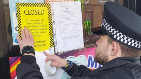 A police officer closing a store in Peterborough. He is putting up a yellow sign, next to another document tapped to the window. The police officer is male, wearing a police hat, he has dark short hair, a beard, has a black watch on his left wrist, and is holding tape in his right hand.