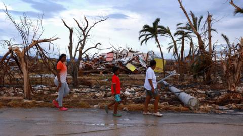 Three people walking past broken trees and blown down houses left by the passage of hurricane Melissa.
