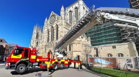 A fire engine with an aerial platform is stationed outside York Minster on a sunny day.