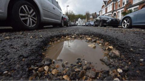 A pothole in the middle of a road with dozens of cars and houses on each side.