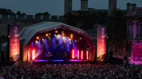 A night-time view of a performance at Lincoln Castle. A large crowd is illuminated by red and pink dazzling neon lights. They are in front of the stage. Two performers are lit by a hazy purple light. 