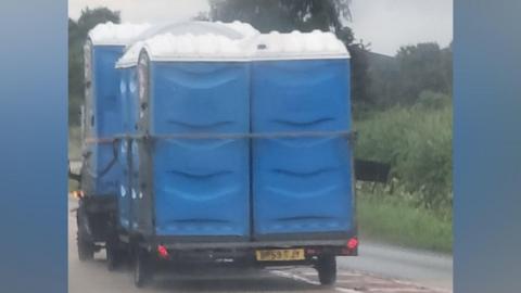 Multiple blue plastic portable toilets are driven around on the back of a trailer.