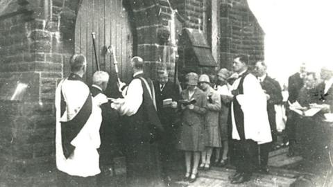 The Bishop of Blackburn at the doors of the new church of St Stephen's Grane after its move in 1927 to officially open it with other church officials and parishioners standing behind him reading a book during the service