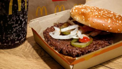 A McDonald's Quarter Pounder in a box next to a plastic cup filled with Coca Cola. The burger has two buns, one of which is seeded, and it has a beef patty, slivered onions, and gherkins in it.