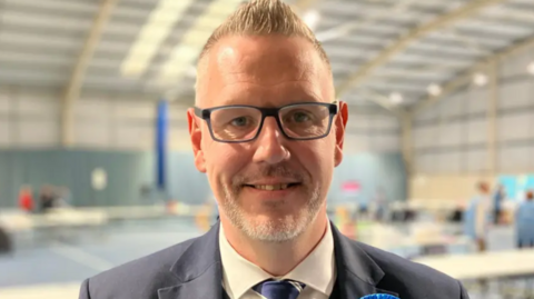 A close-up image of a man with short blonde hair who is wearing dark-framed glasses, a cream-coloured shirt and blue tie and a grey suit. The room behind him is blurred.