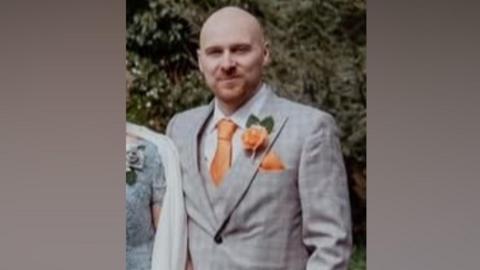 Gareth Underwood, who is bald with a light brown beard. He is wearing a smart light grey suit with an orange tie with matching handkerchief and flower in the lapel