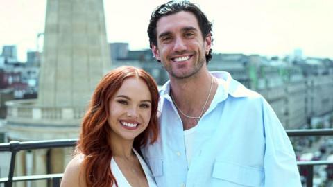 Alexis Warr, a woman in a white top with long, straight red hair, and Julian Caillon, a man in a blue shirt with dark hair slicked back, stand smiling on a balcony with a view of a street with cars and buildings