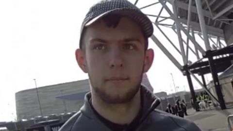 A man with short dark hair and a beard in a grey top and grey cap in front of a large grey building and the corner of a football stand