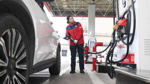 A woman in a red jacket looks at the metre on the petrol pump as she holds the hose near the rear of a white car
