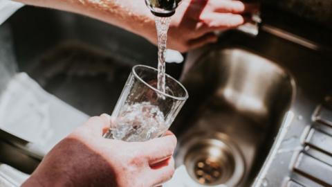 A man is holding a glass underneath a silver tap which is running water. The tap is part of a silver sink which has a draining board on the right.