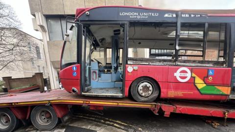 The side of the abandoned red bus, sitting on the bed of a truck as it is removed from the courtyard. The bus is a London United vehicle, with the door missing and all the windows smashed. The floor inside is filthy and the interior is painted a light blue.