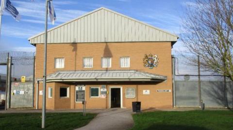 The facade of a two storey prison building seen front on, with fencing and barbed wire either side