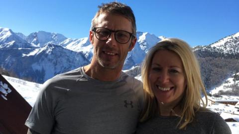 A man with short hair and round glasses, wearing a grey t shirt, smiling next to a woman with blonde hair. Snowy mountains can be seen behind them.