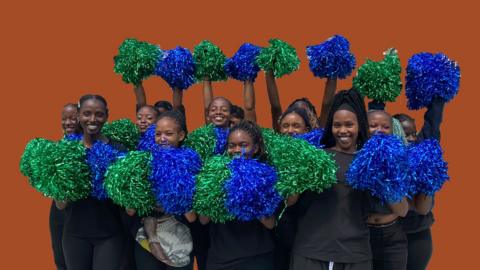 A group of female cheerleaders dressed in black stand and hold up green and blue pom poms