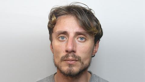 A mugshot photo of a man with a light darkly-coloured beard and dark hair